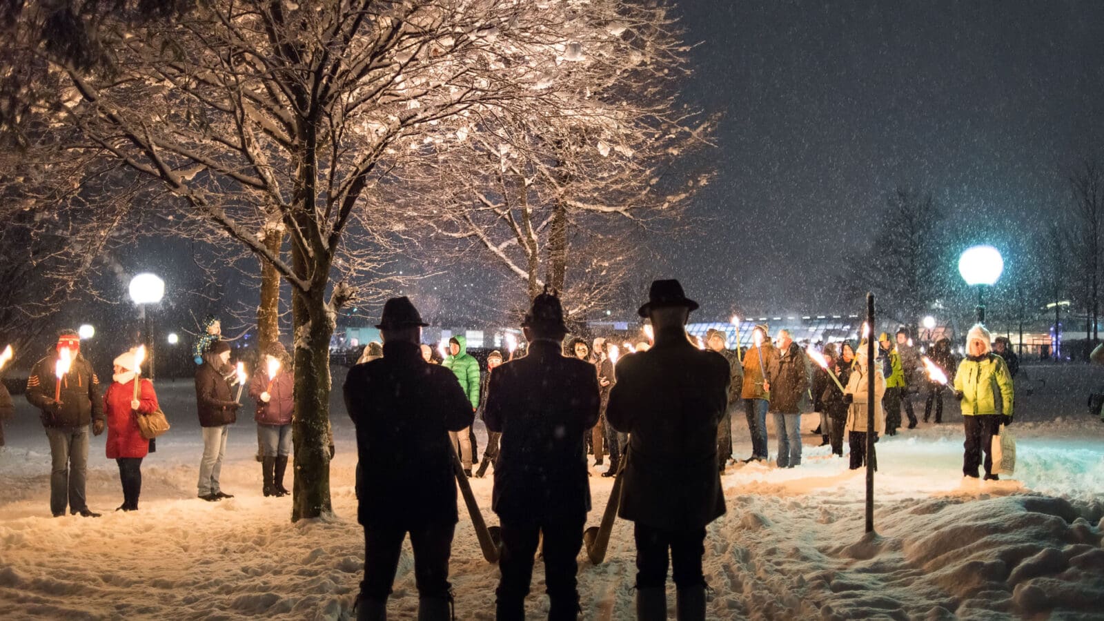 Eine Gruppe von Menschen steht nachts im Schnee in der Nähe von Das Gxund in Bad Hofgastein und hält Fackeln unter schneebedeckten Bäumen. Im Vordergrund stehen drei dunkel gekleidete Gestalten mit Hüten, die sich der vom Fackellicht beleuchteten Gruppe zuwenden.