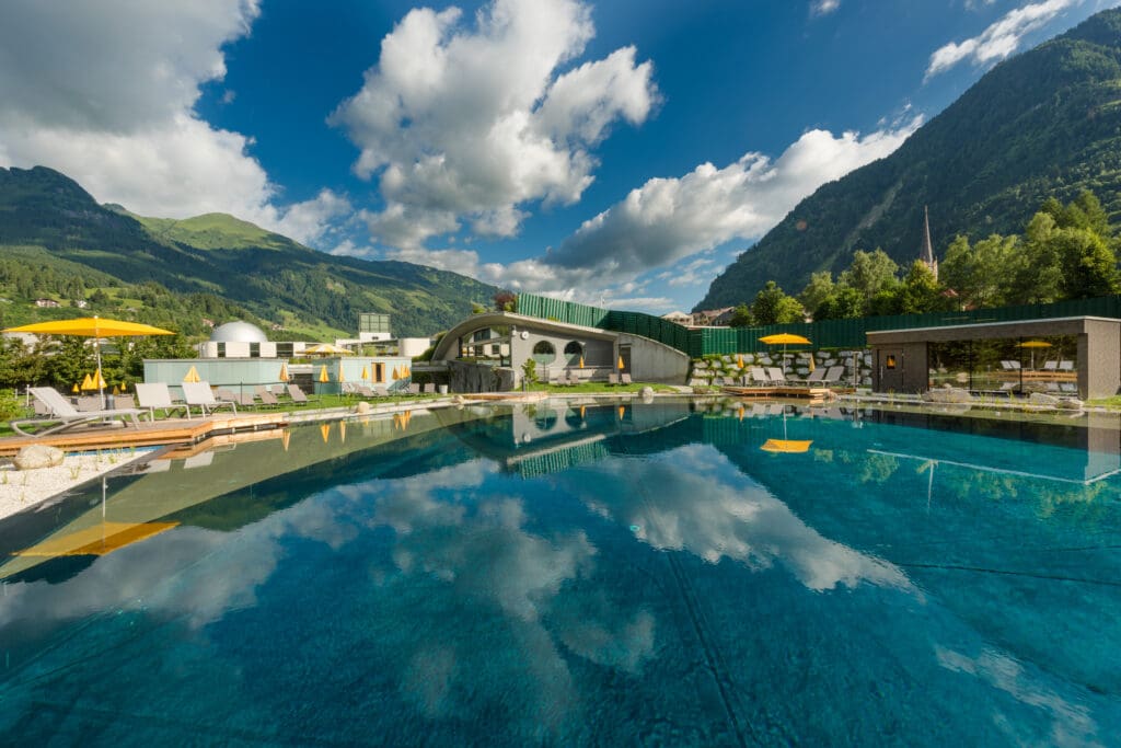 Im klaren Außenpool des Gxund spiegeln sich ein modernes Gebäude, Liegestühle und gelbe Sonnenschirme vor dem Hintergrund der grünen Berge von Bad Hofgastein und einem blauen Himmel mit vereinzelten Wolken.