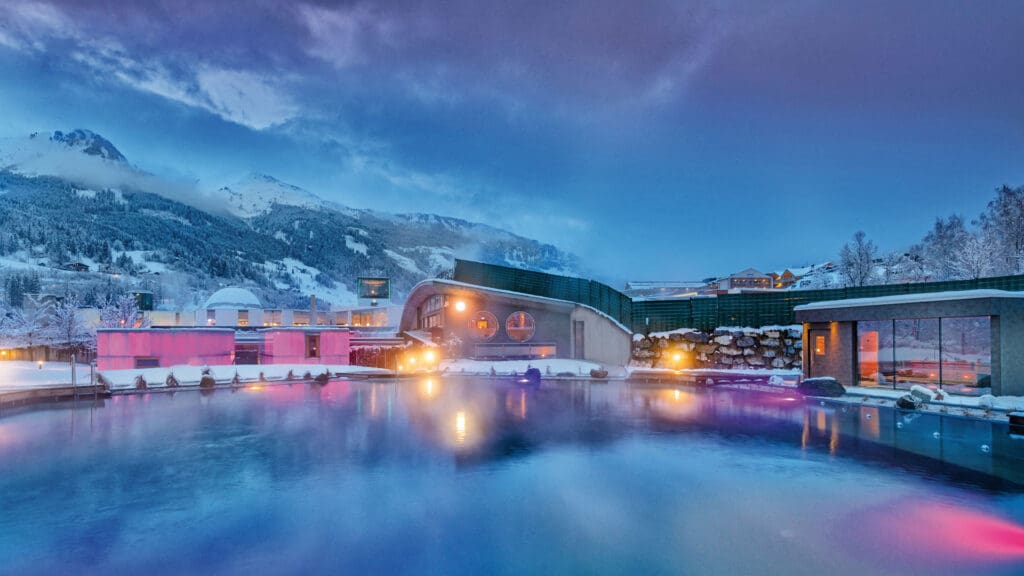 In einem modernen Kurort wie Das Gxund in Bad Hofgastein spiegeln sich leuchtende Lichter in einem dampfenden Außenbecken, das in der Abenddämmerung von Schnee und Bergen umgeben ist und eine ruhige Winteratmosphäre schafft.