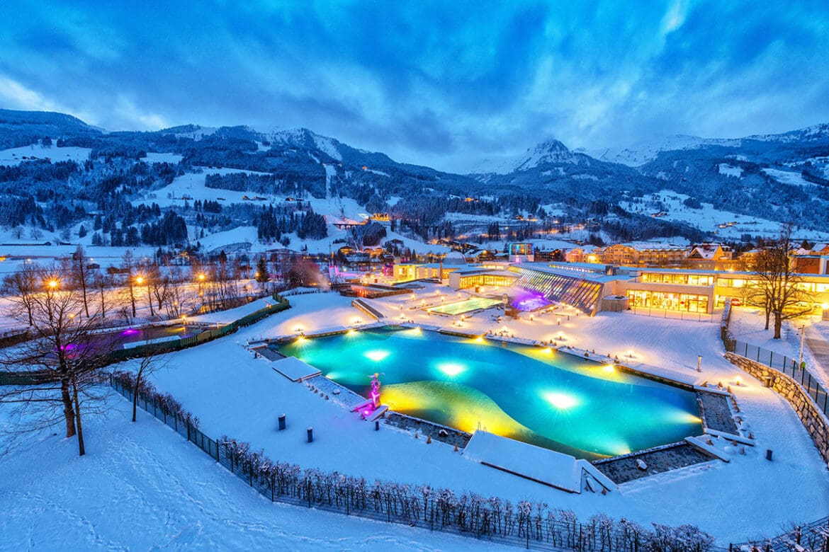 Der Freibadkomplex Das Gxund in Bad Hofgastein erstrahlt in der Abenddämmerung inmitten der verschneiten Berge in bunten Lichtern. Die umliegenden Gebäude und Bäume sind mit Schnee bedeckt, und der Himmel ist blau und wolkenverhangen.