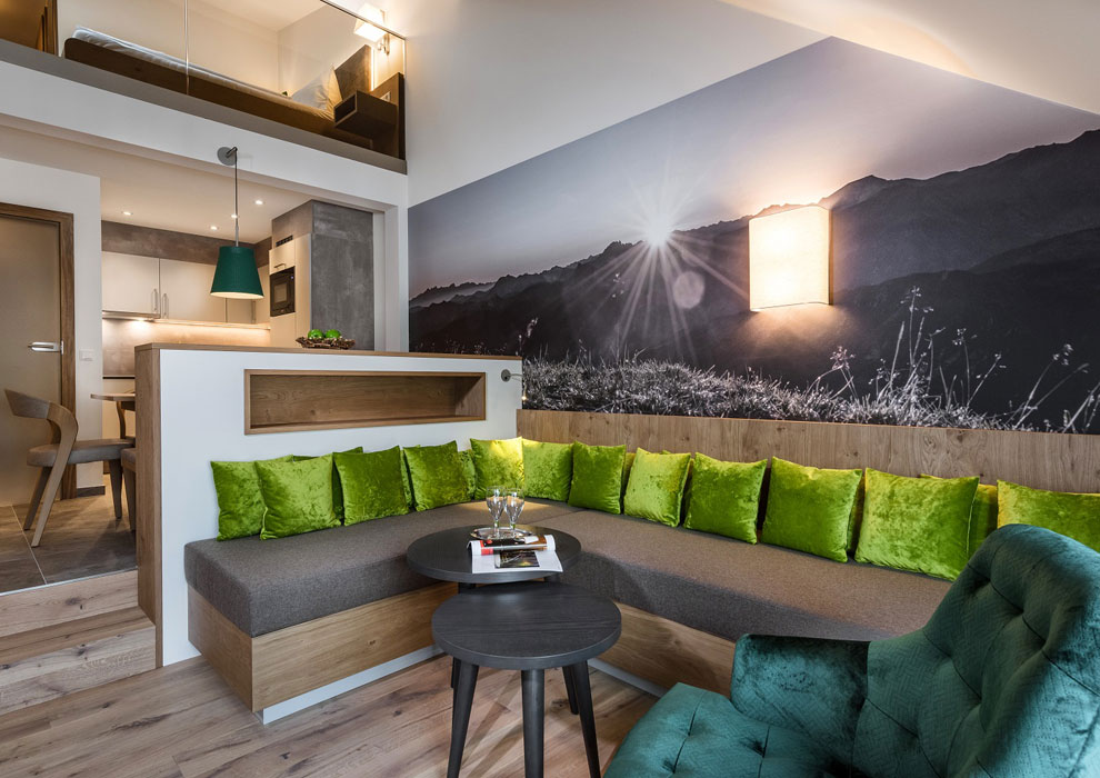 Modernes Wohnzimmer im Das Gxund in Bad Hofgastein mit einem Sektionssofa mit hellgrünen Kissen, runden schwarzen Couchtischen, einem grünen Sessel und einem großen Wandgemälde mit einer sonnenbeschienenen Berglandschaft. Oben ist der Hochbettbereich zu sehen.