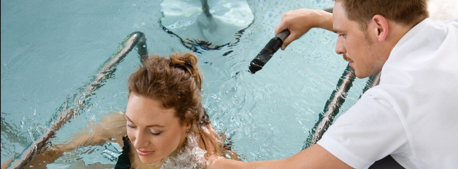 Eine Frau in einem schwarzen Badeanzug sitzt in einem Pool, während eine Person mit einem Handgerät Wasser auf ihre Schulter sprüht, was auf eine Hydrotherapie- oder Radontherapie-Sitzung im Wasser hindeutet.