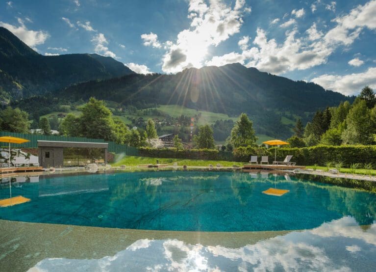 Das Gxund in Bad Hofgastein verfügt über einen runden Außenpool mit Liegestühlen und gelben Sonnenschirmen, umgeben von üppigem Grün und Bergen bei teilweise bewölktem Himmel, wobei das Sonnenlicht auf die Gipfel fällt.