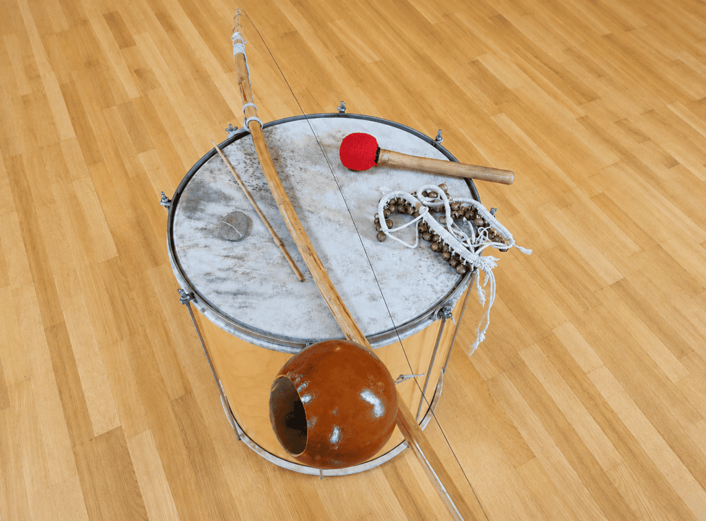 Aktivitäten im Hotel - Taketina Workshop Eine Trommel, ein Berimbau, ein Caxixi-Rüttelinstrument und ein Trommelstock mit roter Spitze liegen auf einem hellen Holzboden – perfekte Instrumente für Spaß und Erlebnisse während Ihres Aufenthalts im Taketina Hotel Gxund Bad Hofgastein – Aktivitäten im Gesundheitsresort.