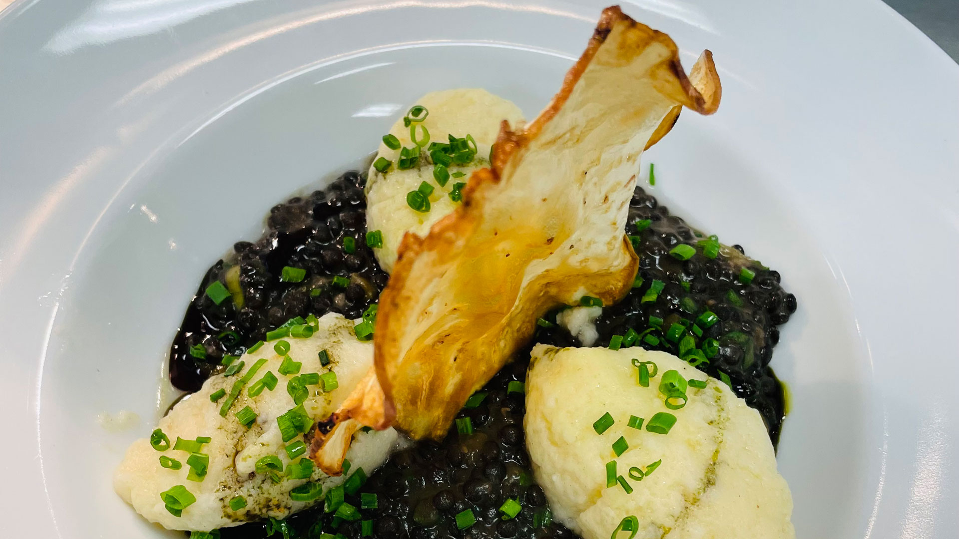 Flaumige Nockerl mit Linsenragout 1 Ein Gourmet-Gericht aus dem Gxund in Bad Hofgastein mit drei Quenelles aus cremiger Mousse, schwarzen Linsen, fein geschnittenem Schnittlauch und einer knackigen Gemüsechip-Garnitur, elegant serviert auf einem weißen Teller.