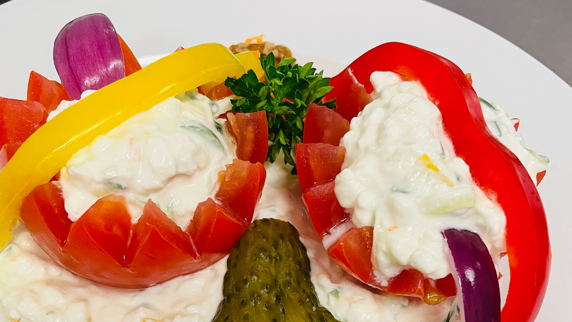 Nahaufnahme eines bunten Salats im Das Gxund in Bad Hofgastein: Tomaten- und Paprikaschalen, gefüllt mit cremigem Hüttenkäse, garniert mit Petersilie, roten Zwiebeln und einer Gurke auf einem weißen Teller.