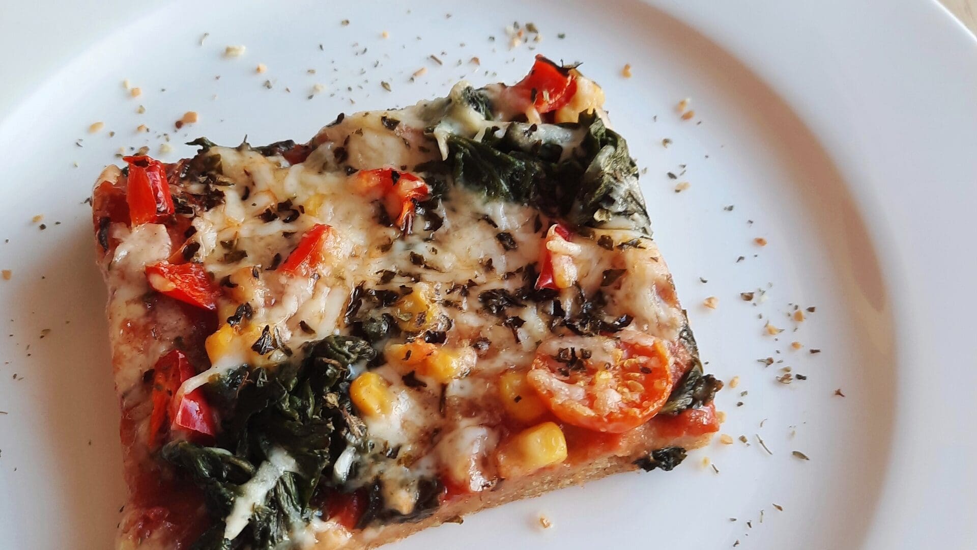 Kartoffelpizza 1 Ein quadratisches Stück Gemüsepizza von Das Gxund in Bad Hofgastein, belegt mit geschmolzenem Käse, Spinat, rotem Paprika und Kräutern auf einem weißen Teller. Im Hintergrund sorgen Kirschtomaten und Kräuter für Farbe.