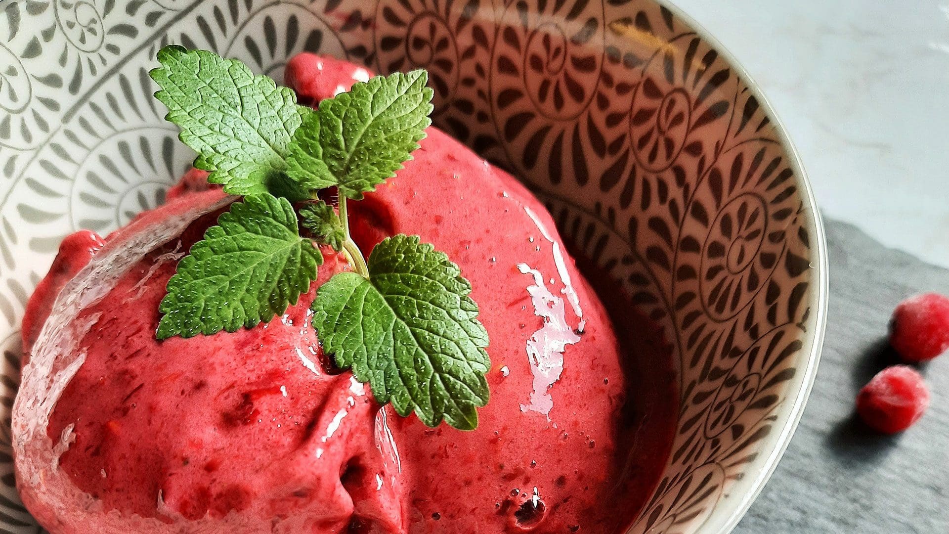 Eine Schale mit leuchtend rotem Beeren-Sorbet wird im Das Gxund, Bad Hofgastein, mit frischen Minzblättern garniert. Die kunstvoll gemusterte Schale enthält eine erfrischende Leckerei, ein paar Beeren sind kunstvoll an der Seite platziert.