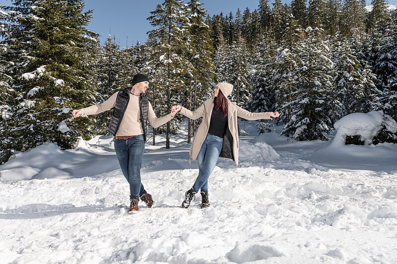 Ein Mann und eine Frau halten sich an den Händen und spazieren fröhlich durch den tiefen Schnee in einem Wald, umgeben von schneebedeckten Bäumen unter einem klaren blauen Himmel. Sie tragen Winterkleidung wie Mäntel, Mützen und Stiefel.