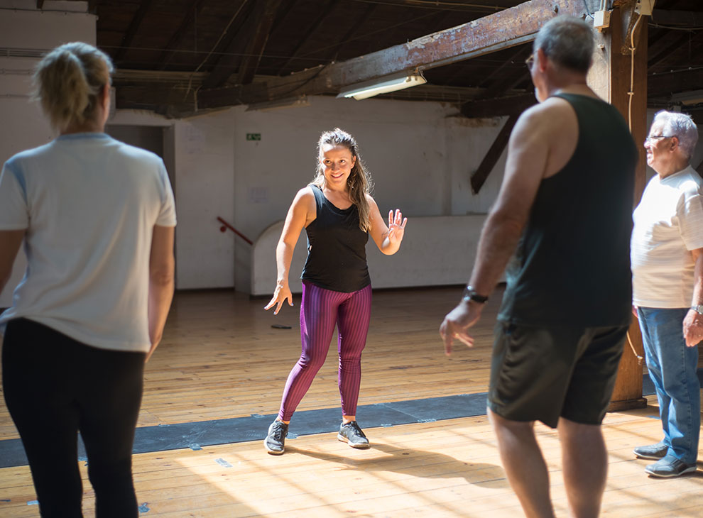 Aktivitäten im GXUND - Line Dance Im Gxund in Bad Hofgastein leitet eine Frau in Sportkleidung einen Gruppenfitnesskurs in einem sonnendurchfluteten, großen Raum mit Holzboden. Drei Erwachsene stehen ihr gegenüber und nehmen an der Aktivität teil.