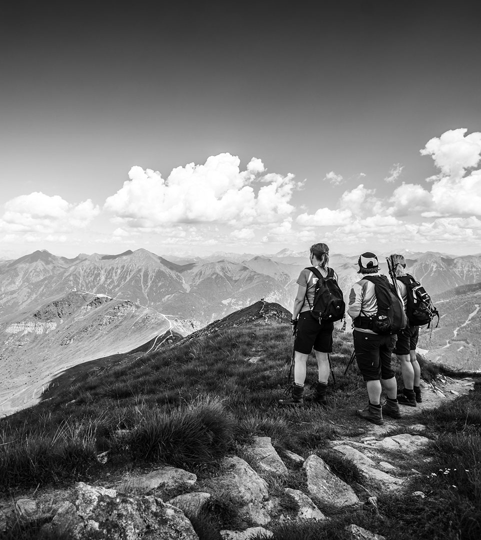 Drei Wanderer mit Rucksäcken stehen auf einem Bergpfad in der Nähe von Bad Hofgastein und blicken auf ferne Gipfel und Wolken unter einem klaren Himmel; das Schwarz-Weiß-Bild fängt die landschaftliche Schönheit rund um Das Gxund ein.