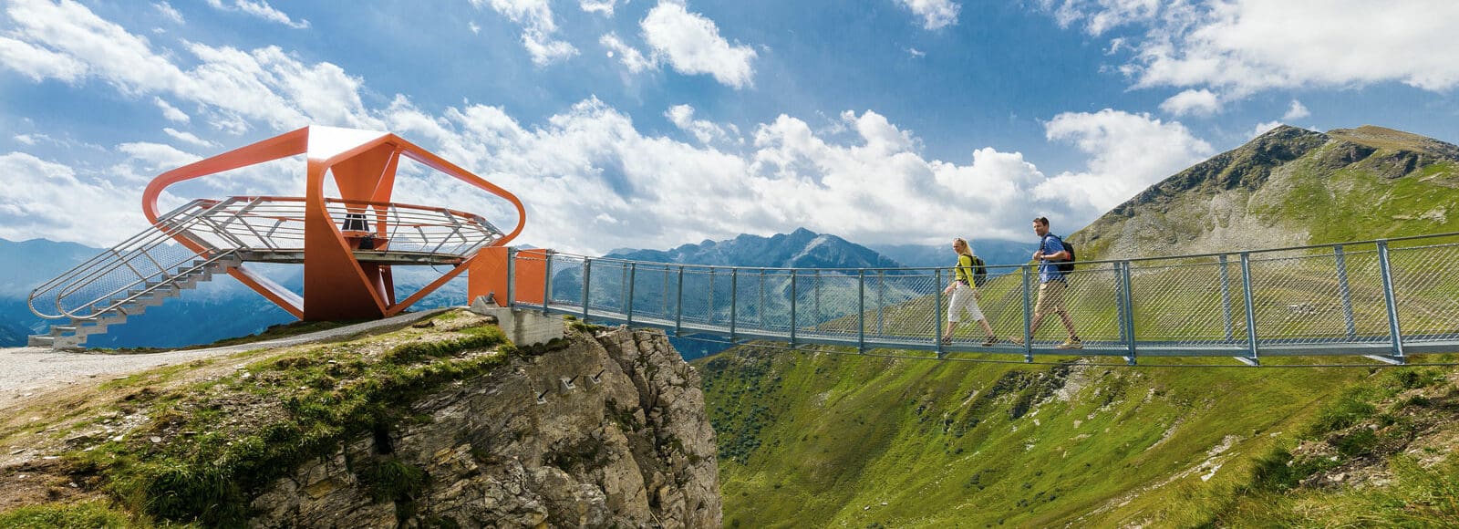 Zwei Personen gehen über eine metallene Hängebrücke über ein tiefes Bergtal in der Nähe von Bad Hofgastein zu einer modernen, orangefarbenen Aussichtsplattform mit Blick auf grüne Berge unter einem teilweise bewölkten Himmel.