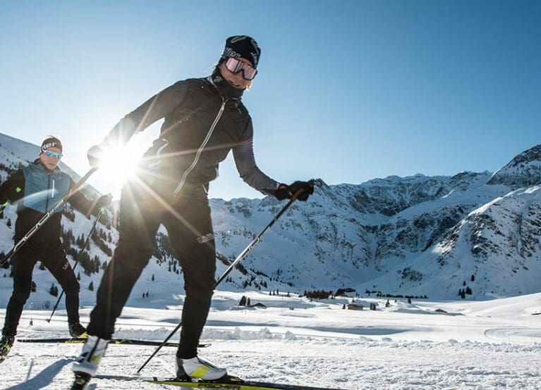 Zwei Langläufer in einer verschneiten Berglandschaft bei Bad Hofgastein an einem sonnigen Tag, mit hellem Sonnenlicht im Rücken und den majestätischen Gipfeln um Das Gxund im Hintergrund.