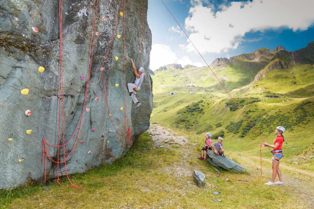 Shooting Gasteiner Bergbahnen Sommer 2013 Eine Person klettert eine Felswand mit bunten Griffen im Freien in Bad Hofgastein, während eine andere sie sichert. Zwei Kinder, die Das Gxund besuchen, sitzen auf einem Felsen in der Nähe und beobachten die grasbewachsene, bergige Landschaft unter einem teilweise bewölkten Himmel.