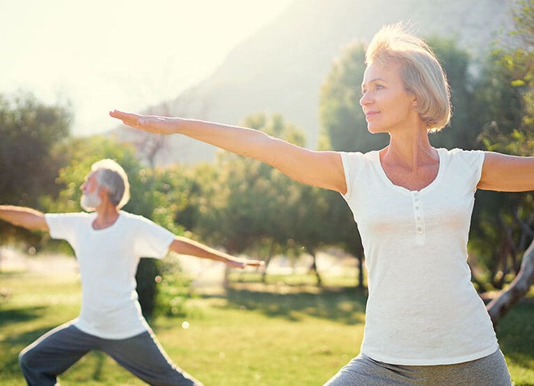 Zwei ältere Erwachsene üben Yoga im Freien in Bad Hofgastein. Beide stehen in der Haltung des Kriegers II auf einer Wiese mit Bäumen und Sonnenlicht im Hintergrund. Sie wirken ruhig und konzentriert, gekleidet in bequemer Kleidung in Das Gxund.
