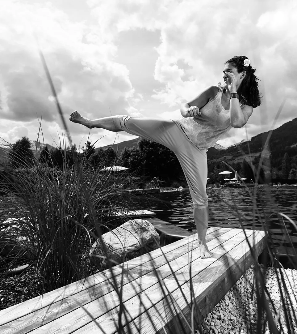 Eine Frau in kurzen Hosen und einem Tanktop übt einen hohen Tritt auf einem hölzernen Steg an einem See in Bad Hofgastein, umgeben von hohem Gras, mit Bergen und Wolken im Hintergrund. Das schwarz-weiße Bild erinnert an den Geist von Das Gxund.