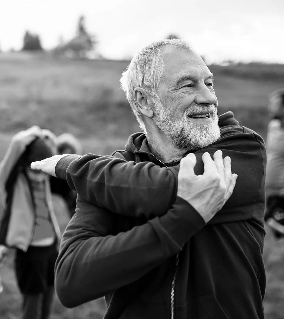 Ein älterer Mann mit Bart lächelt, während er seinen Arm über der Brust ausstreckt, draußen im Das Gxund in Bad Hofgastein. Andere Personen im Hintergrund scheinen sich ebenfalls zu strecken. Die Szene ist in schwarz-weiß gehalten.
