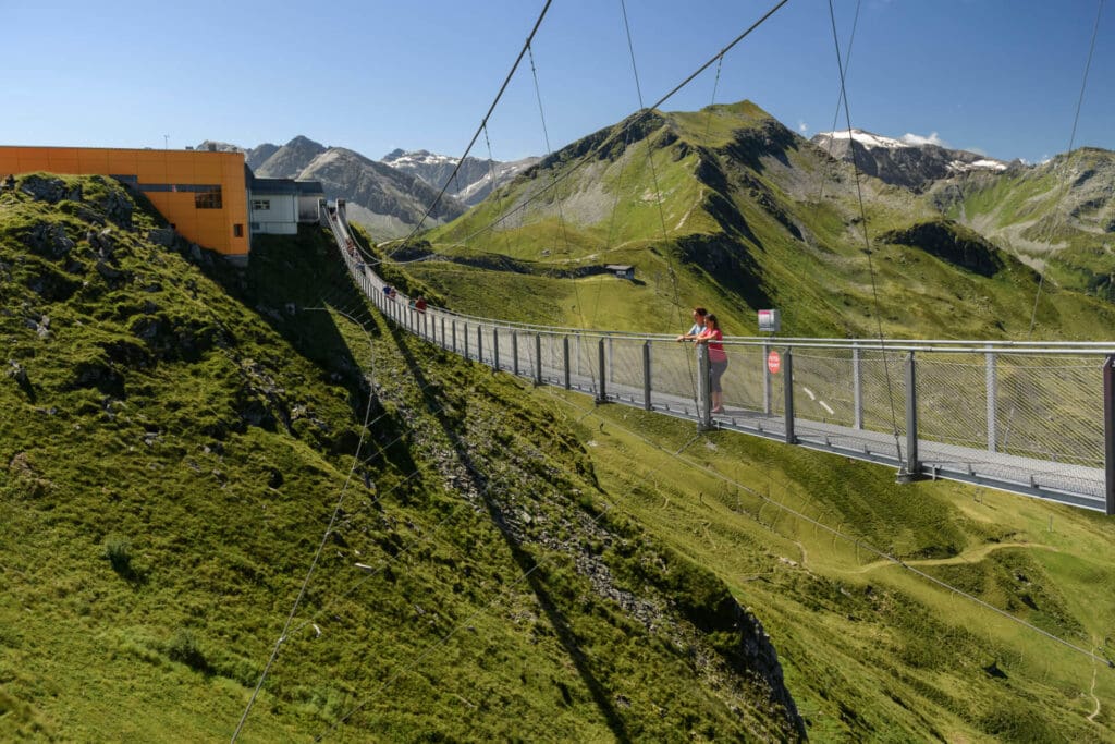 Gasteiner Bergbahnen Hängebrücke Eine Person steht auf einer langen Hängebrücke über einem tiefgrünen Tal in Bad Hofgastein, umgeben von Bergen unter einem klaren blauen Himmel, mit dem gelben Gebäude Das Gxund an einem Ende der Brücke.