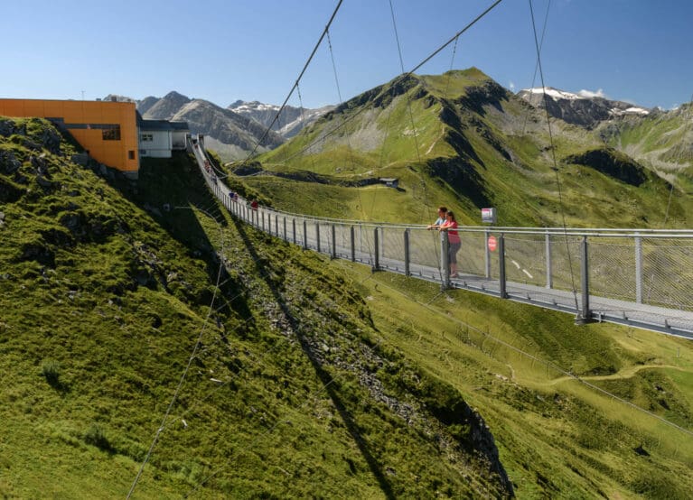 Eine Person steht auf einer langen Hängebrücke über einem tiefgrünen Tal in Bad Hofgastein, umgeben von Bergen unter einem klaren blauen Himmel, mit dem gelben Gebäude Das Gxund an einem Ende der Brücke.