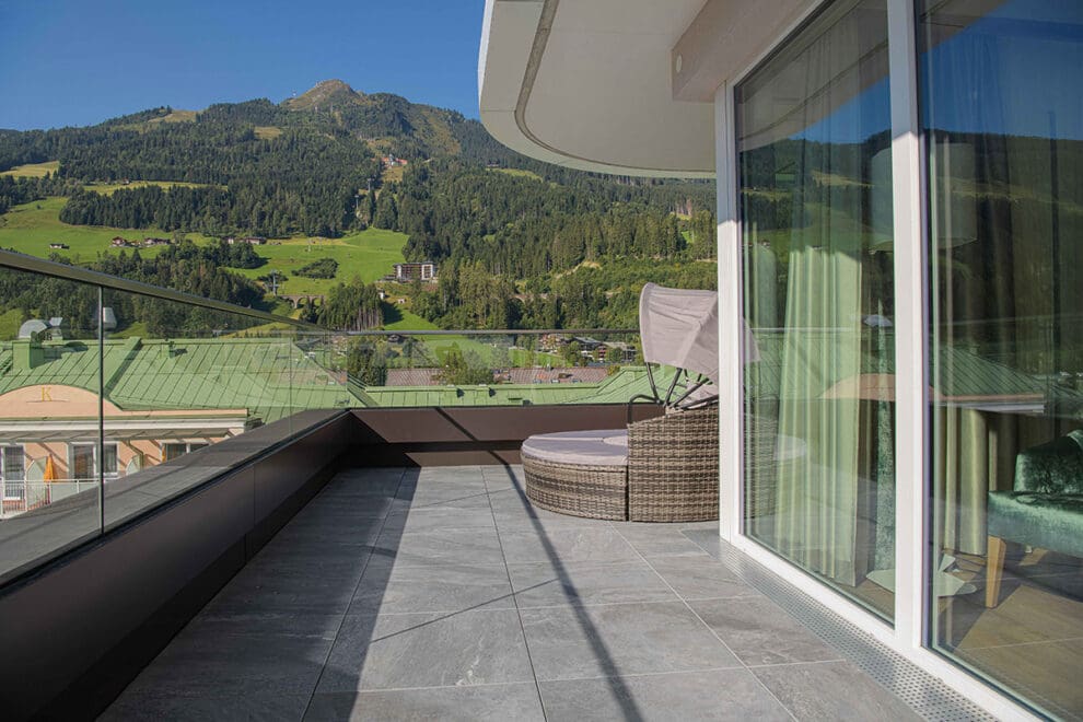 Ein moderner Balkon im Das Gxund in Bad Hofgastein ist mit grauen Fliesen und einem Glasgeländer ausgestattet und bietet einen Blick auf grüne Hügel und Berge unter einem klaren blauen Himmel. Neben den Glastüren steht ein runder Korbsessel.