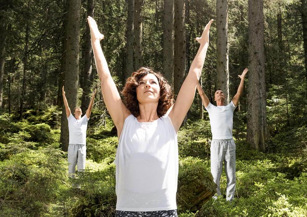 Yoga in Bad Hof Gastein - Aktiv anders Drei Personen stehen mit erhobenen Armen in einem Wald in der Nähe von Bad Hofgastein und scheinen Yoga oder Dehnungsübungen zu machen. Das Sonnenlicht fällt durch die hohen Bäume und schafft die ruhige, natürliche Atmosphäre von Das Gxund.